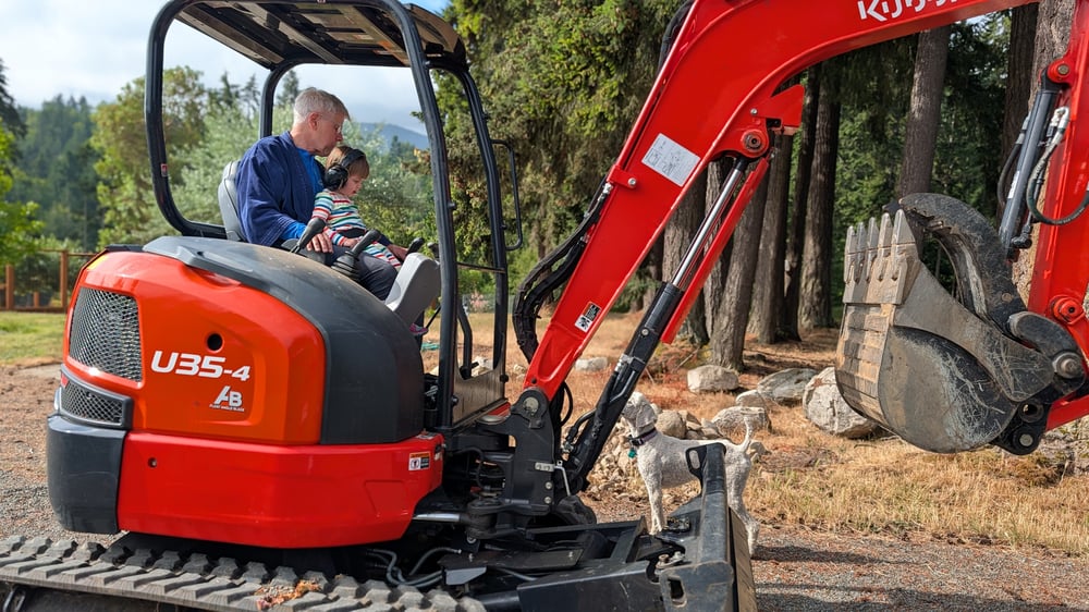 Steve Shafer driving a bulldozer with his grandson Asa, age 2.